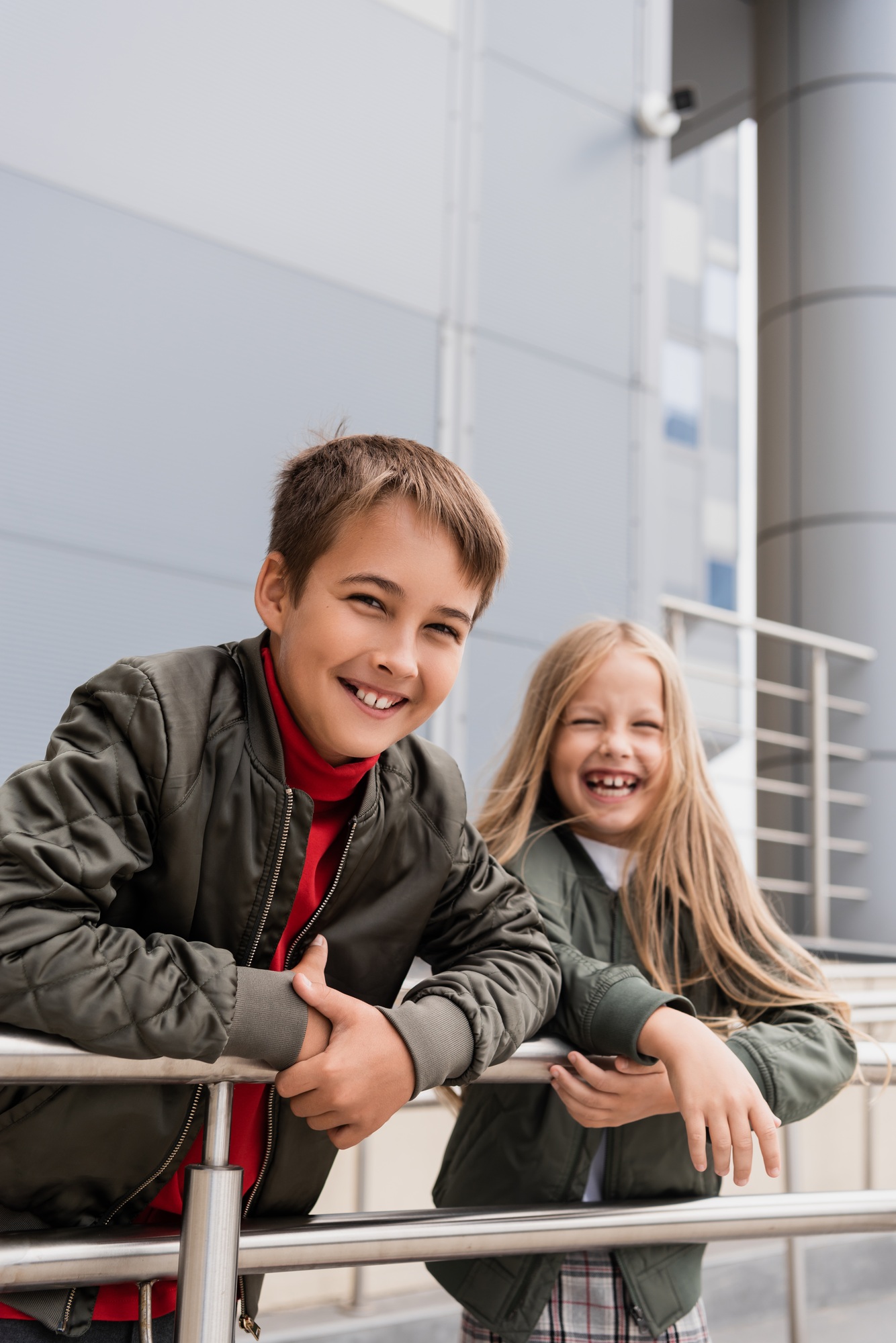cheerful preteen kids in bomber jackets leaning on metallic handrails near mall