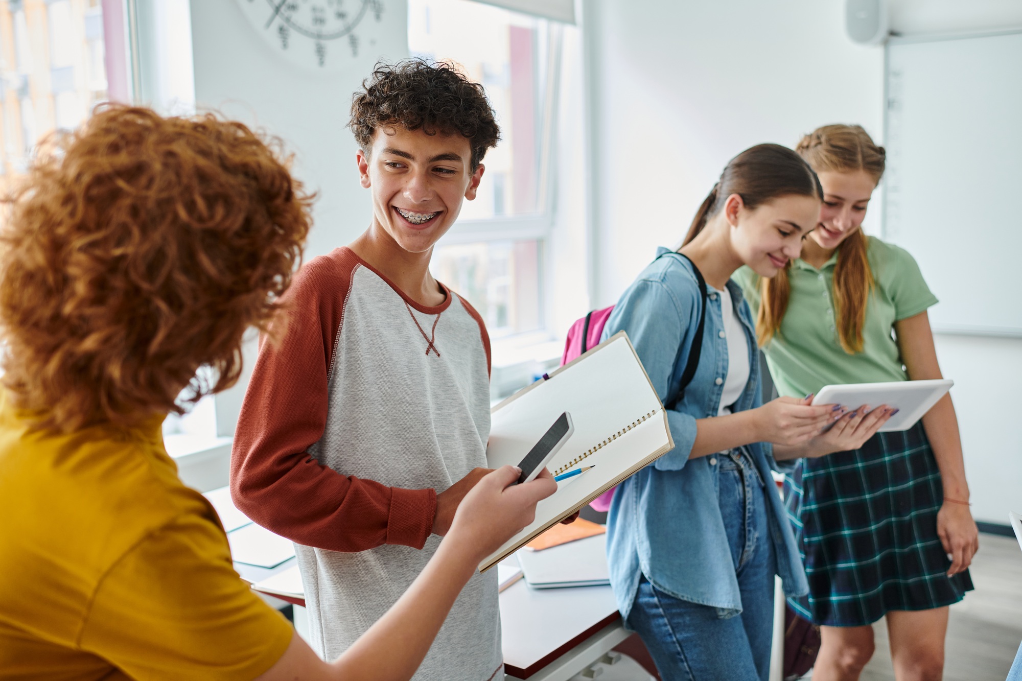 Smiling schoolboy with braces holding notebook near friends with devices in classroom in school